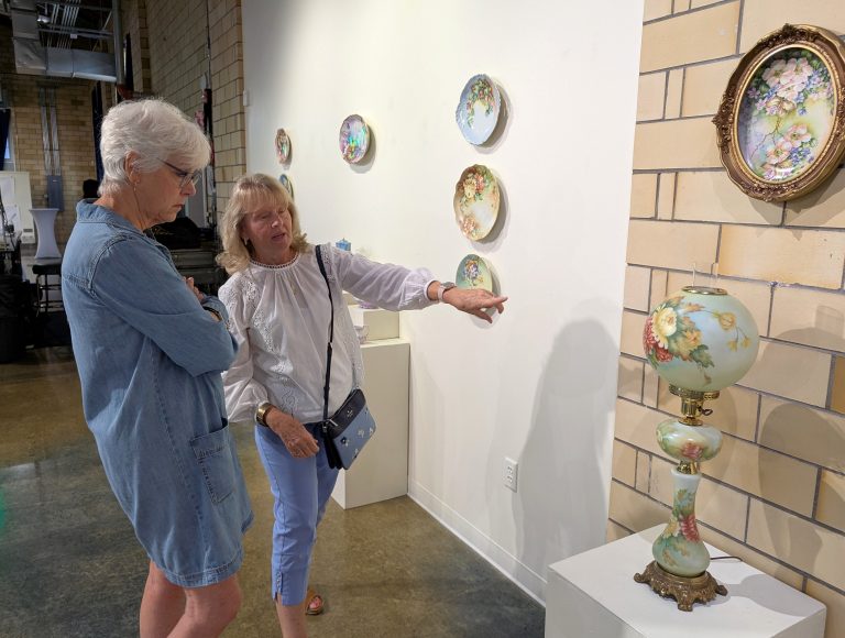 Wassenberg Art Center’s Jayne Smith and Sherry Rager discuss Rager’s grandmother’s porcelain art which is on display at the center through August. (Sam Shriver/CherryRoad Media)
